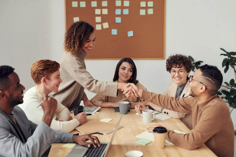 Drie mannen en drie vrouwen zitten samen aan een vergadertafel. Een man en een vrouw tegenover elkaar geven elkaar een hand.