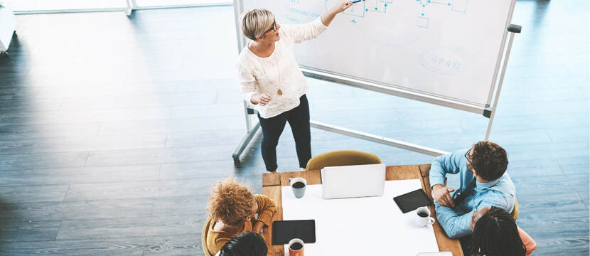 Vier personen zitten aan een vergadertafel met daarvoor een vrouw die uitleg geeft over een tekening op een whiteboard.